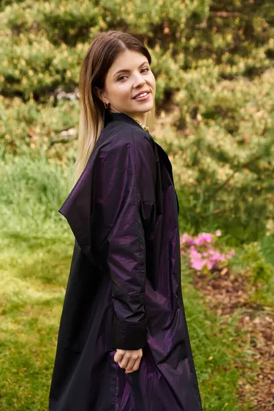 Koyu renk ceketli genç bir kadın yemyeşil bahçeye bakıyor..