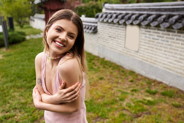 A happy young woman in a pink dress embraces herself for warmth.
