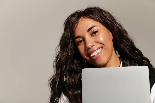 A young woman smiles brightly while holding a laptop against her chest in a studio setting.