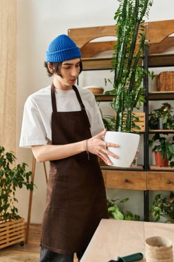 Önlüklü genç adam dikkatle bir bitkiyi sıcak bir stüdyoda daha büyük bir kaba taşıyor..