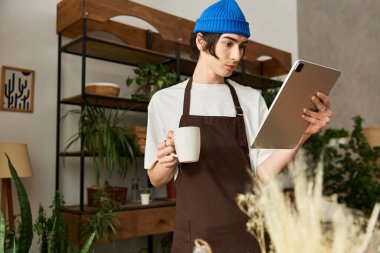 Stüdyoda elinde kahve fincanı ve tablet tutarken çiçeklerle ilgilenen yakışıklı genç adam..