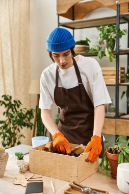 Kahverengi önlüklü ve mavi bereli genç bir adam canlı bir stüdyoda bitki naklediyor..