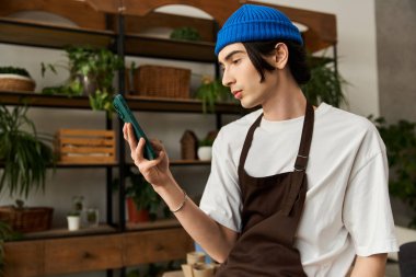 Kahverengi önlüklü genç bir adam telefonuna odaklanırken bitki naklediyor..