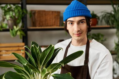 Mavi bereli genç bir adam yeşil, sıcacık bir stüdyoda çiçek tutuyor..