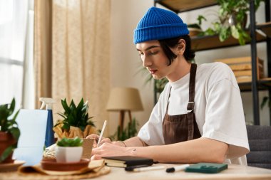 Genç bir adam parlak, rahat bir stüdyoda canlı bitkilerin arasında deftere bir şeyler yazıyor..