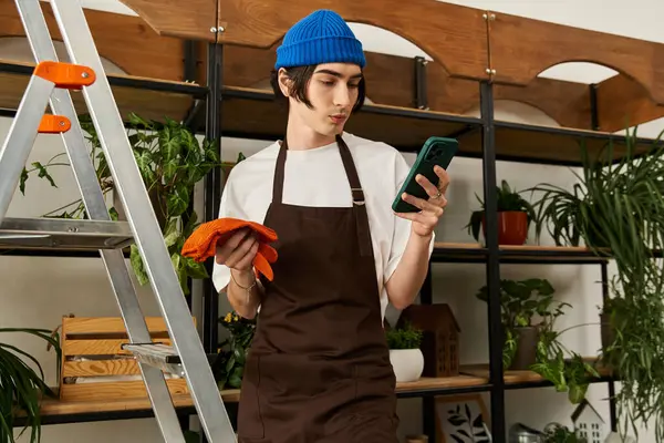 Şık önlüklü genç adam, bir stüdyoda yemyeşil bitkilerle çevriliyken telefonunu kullanıyor..