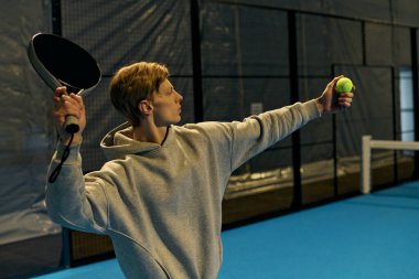 Genç bir adam, kapalı bir kort ortamında yoğun bir Padel maçı sırasında hizmet etmeye hazırlanıyor..