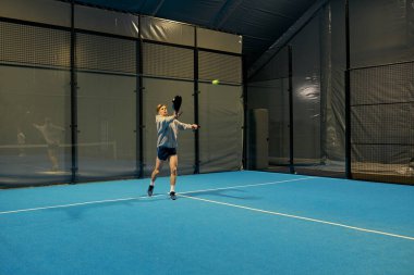 Kendini işine adamış genç bir adam, bir sonraki atışına odaklanarak, kapalı bir padel kortunda kararlılıkla antrenman yapıyor..