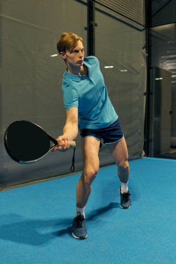 Genç bir adam modern bir sahada Padel oynarken atletizmini sergiliyor..