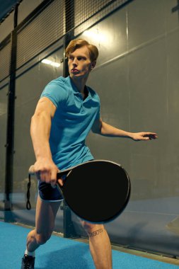 Atletik giysiler içindeki genç bir adam evde Padel oynarken odaklandığını ve çevikliğini gösteriyor..