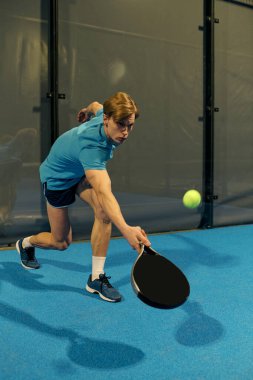 Sporcu bir genç parlak renkli bir kortta padel topuna vurmak için hamleler yapar..