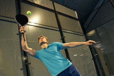 Genç bir sporcu, Padel sahasında atletik yeteneklerini sergileyerek topa hizmet etmeye hazırlanıyor..