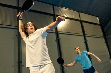 İki genç adam kapalı sahada maharetlerini ve takım çalışmalarını sergileyerek padel oynuyor..
