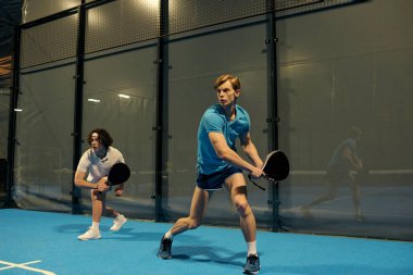 Modern bir mekanda Padel oynarken atletizm sergileyen iki genç adam..