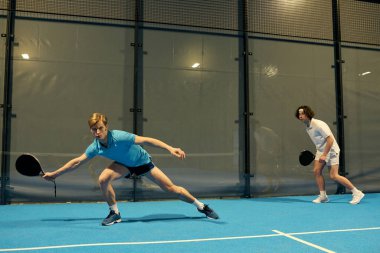 İki genç adam, açık mavi bir sahada padel oynarken yeteneklerini sergiliyorlar..