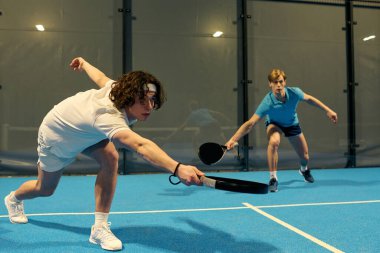 İki genç adam yeteneklerini açık mavi bir sahada rekabetçi bir padel oyununda sergiliyor..
