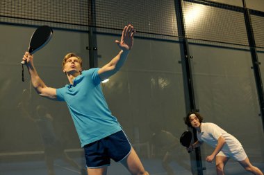 İki genç sporcu, rekabetçi bir maç sırasında içeride Padel oynarken yeteneklerini sergiliyorlar..