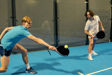 İki genç adam kapalı bir sahada Padel oynarken yeteneklerini sergiliyorlar..