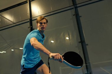 Genç bir atlet Padel oynarken yoğun bir şekilde odaklanır, raketini ustalıkla kullanır..