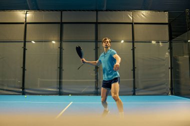 Sportif giyimli genç bir adam canlı bir mavi kortta hızlı bir Padel oyununa ustalıkla katılıyor..