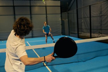 İki genç adam kapalı bir sahada padel oynarken yeteneklerini sergiliyorlar..