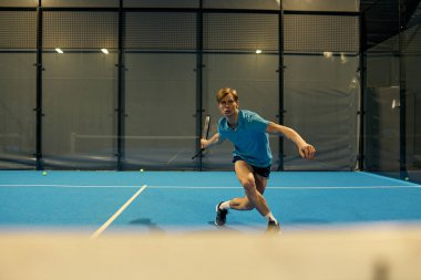 Spor giyimli genç bir adam Padel antrenmanı yapıyor, çevikliğini gösteriyor ve mahkemeye odaklanıyor..