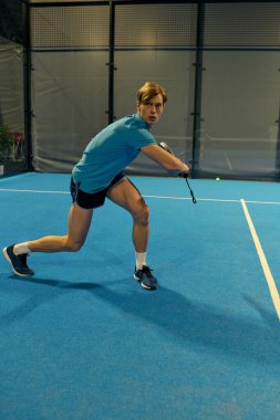 Genç bir adam topa Padel kortunda vurmaya hazırlanırken dikkatle odaklanıyor..