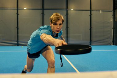 Atletik genç adam Padel oynarken çok güçlü bir atış yapıyor. Önündeki oyuna odaklanıyor..