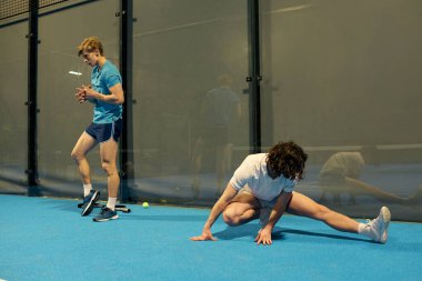 İki genç adam mavi bir padel kortunda sıkı çalışıyor, yeteneklerine ve sağlıklarına odaklanıyor..