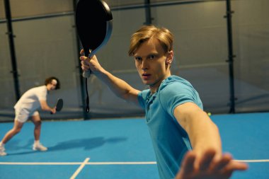 İki genç sporcu, kapalı bir sahada Padel oynarken yeteneklerini sergiliyorlar..