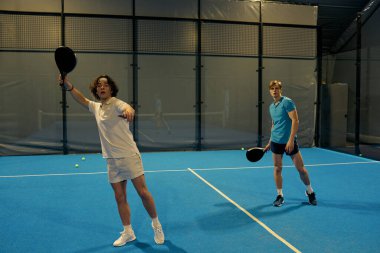 İki genç adam sahada raketlerle padel oynarken yeteneklerini sergiliyorlar..