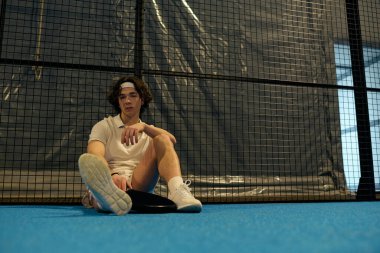 Spor giyim sektöründe genç bir adam maça odaklanmış, mavi padel kortunda oturuyor.