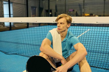 Genç bir atlet padel kortunda dinleniyor, omzuna havlu sarılı, sonraki hamlesini düşünüyor..