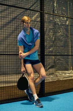 Genç bir adam canlı spor kıyafetleriyle Padel oynarken atletizmini gösteriyor..