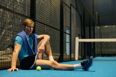 Genç bir sporcu, oynadıktan sonra mavi bir padel kortunda oturmuş atletizmini sergiliyor..