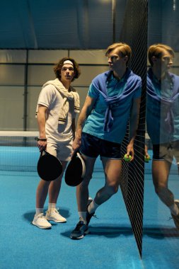 Spor giyimli iki genç, coşkuyla mavi bir sahada Padel maçına hazırlanıyor..