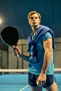 Atletik giyimli genç bir adam kapalı bir padel kortunda harekete geçmeye hazırlanıyor. Yeteneğini sergiliyor..