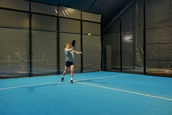 Самоотверженный молодой человек тренируется с решимостью на крытом падель-корте, фокусируясь на своем следующем выстреле.