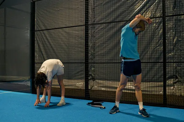 İki genç, mahkeme ağlarının yanında ısınma hareketleri yaparak antrenmana hazırlanıyor..
