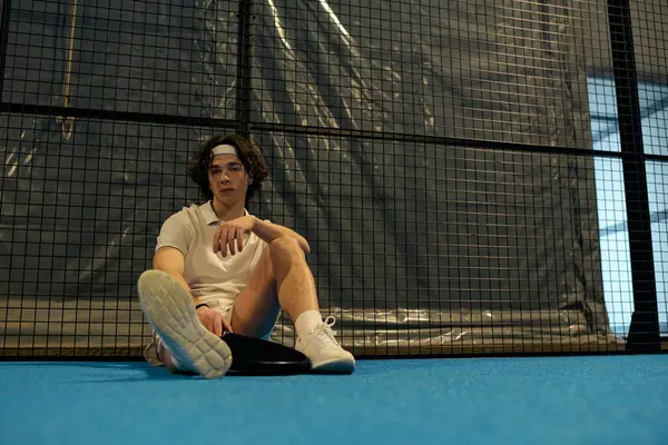 Spor giyim sektöründe genç bir adam maça odaklanmış, mavi padel kortunda oturuyor.