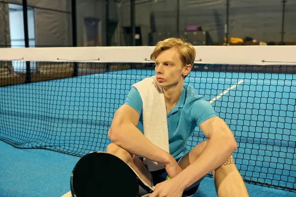 Genç bir atlet padel kortunda dinleniyor, omzuna havlu sarılı, sonraki hamlesini düşünüyor..