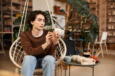 Şık bir genç adam, seramik eserlerini sanatla dolu canlı bir stüdyoda dikkatlice inceliyor..