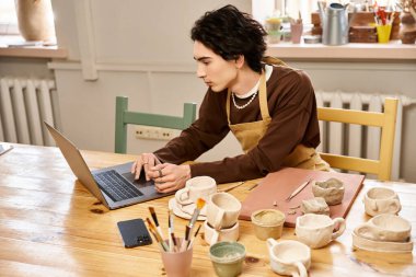 Yaratıcı adam sanatsal bir çalışma alanında dizüstü bilgisayar kullanırken çömlek tasarımlarına odaklanıyor