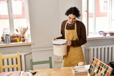 Önlüklü yetenekli bir adam, yaratıcılıkla dolu güneşli bir stüdyoda çömlek malzemeleriyle meşgul oluyor..