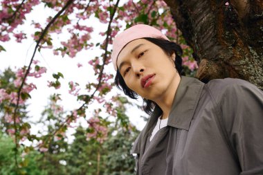 Gri trençkotlu şık bir genç adam kendine güvenen bir kiraz ağacının yanında duruyor..