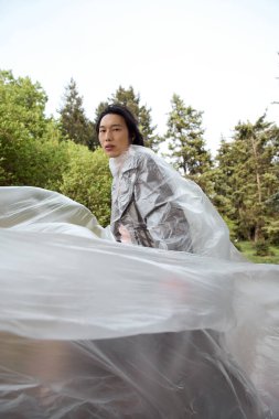 Genç Asyalı bir adam parkta yürür polietilen pozlar verir, sürdürülebilirlik kavramı.