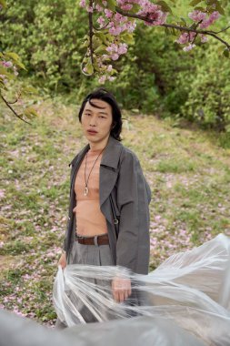 Gri elbiseli genç bir adam zarif bir şekilde parkta duruyor, polietilen ile poz veriyor.