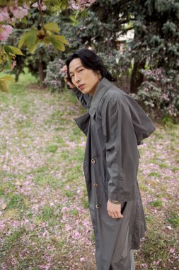 Şık gri bir trençkot giymiş genç bir adam çiçeklerle dolu canlı bir parkta geziniyor..