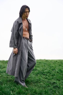 Genç Asyalı bir adam canlı bir parkı keşfederken şık gri kıyafetiyle güvenle yürüyor..