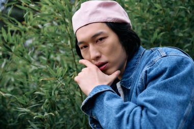 Şık kot elbiseli Asyalı genç bir adam yeşil yaprakların arasında rahatça poz veriyor..